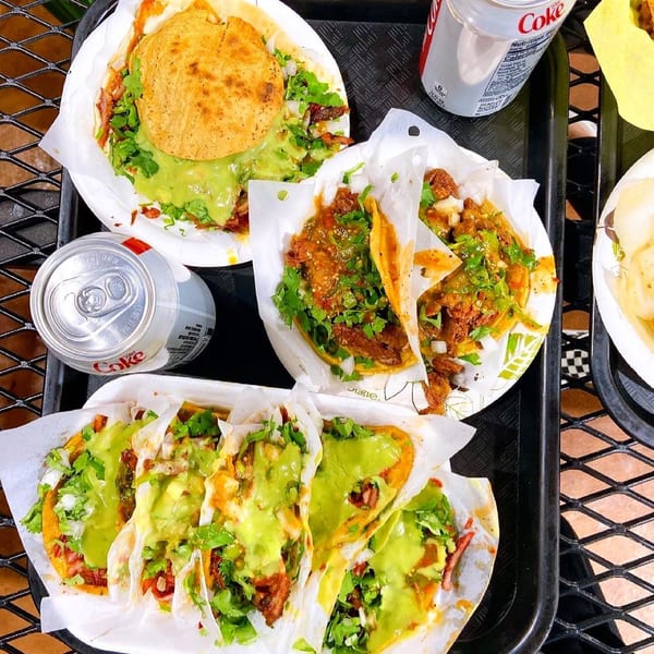 A black tray filled with various Tacos El Pastor tacos and two cans of Diet Coke.