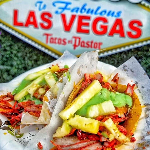 Two Tacos El Pastor with red meat, pineapple, and green sauce, in front of a Las Vegas sign.