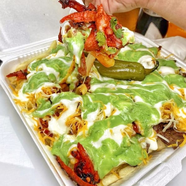 A hand lifts a forkful of Tacos El Pastor's loaded fries with green and white sauce.