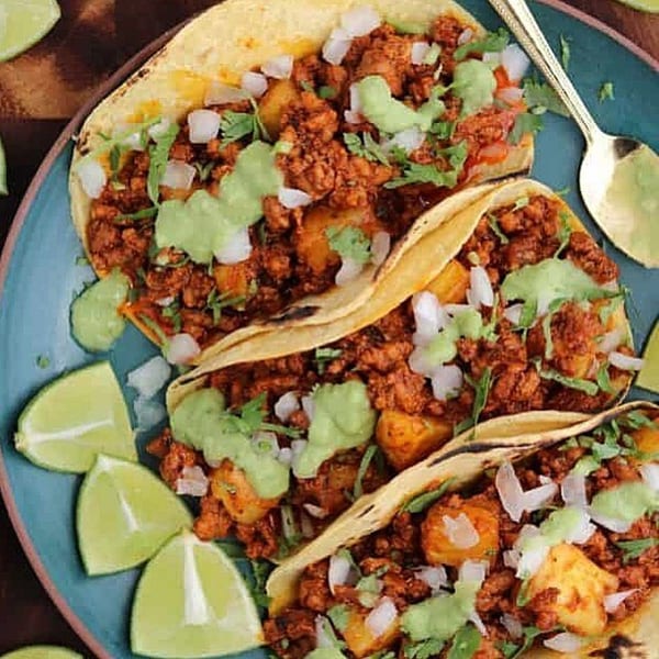 Three Tacos El Pastor on a blue plate, garnished with lime wedges and a golden spoon.