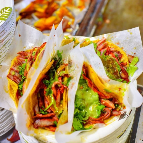 Three Tacos El Pastor, filled with red meat, green salsa, and cilantro, wrapped in paper.