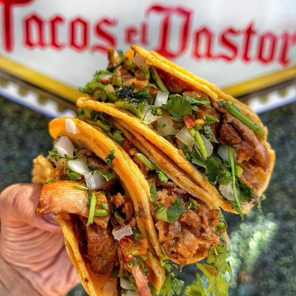 Three Tacos El Pastor tacos, filled with meat, onions, and cilantro, held in a hand.